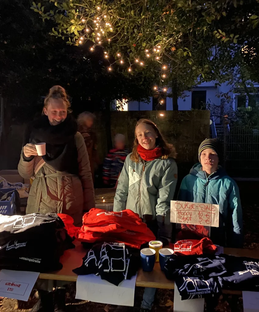 Eine Frau vom Förderverein und zwei Kinder stehen beim Verkaufsstand des Fördervereins beim Lichterfest der Grundschule an der Lessingstrasse Bremen